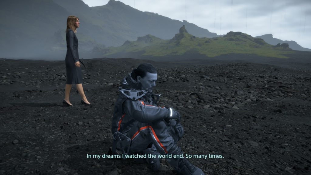 A woman walks on a black-sand beach, around a grayscale, scruffy-looking man in overalls sat in the foreground.