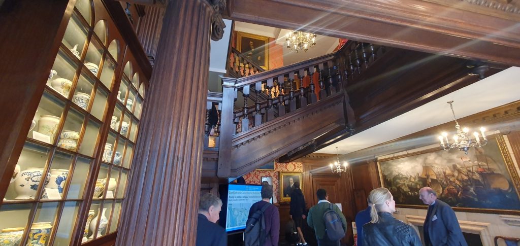 A dark wood staircase climbs from small but well-decorated area, with porcelain in a display case and a painting of a naval  battle on the wall, lit by chandeliers.