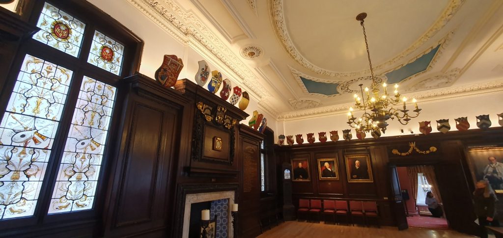 An ornate wood-panelled room with stained glass is decorated with various shields mounted near the tromp-loeil ceiling, where a brass chandelier bathes the paintings of the room in a warm light.