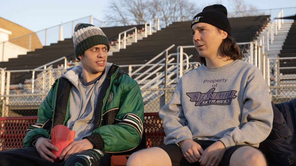 Pete Davidson and Paul Dano sit on a bench at an athletics track and talk.