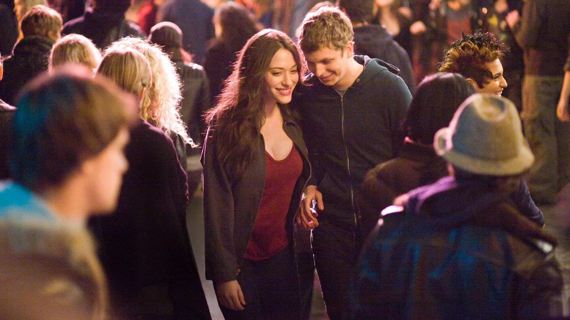 Kat Dennings and Michael Cera listen to shared headphones in a crowded room.