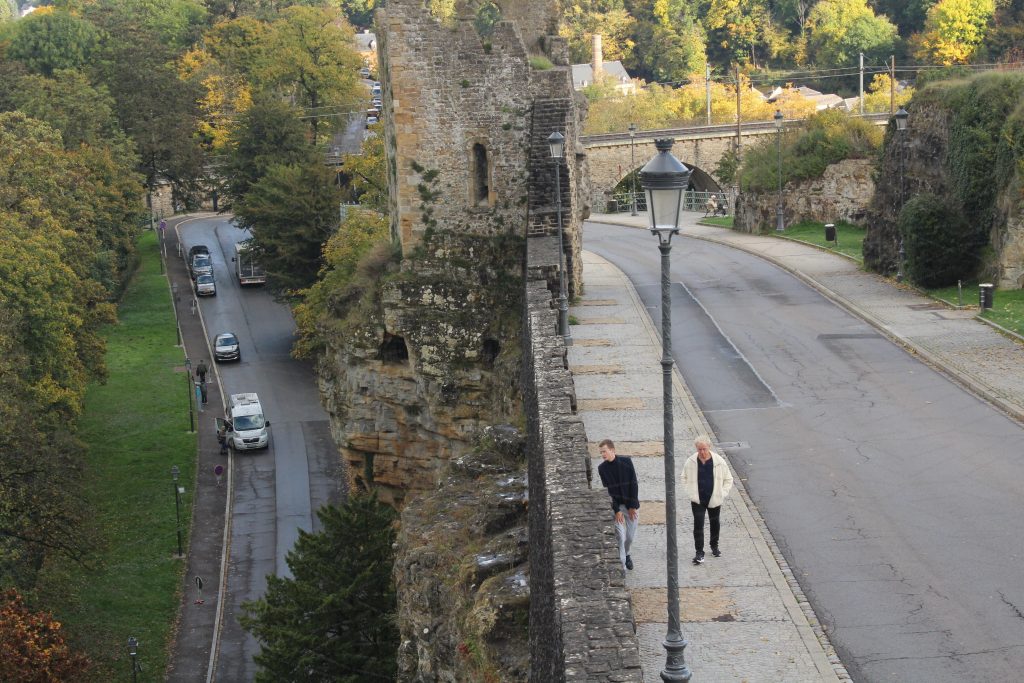 A man peers over an aged wall that forms part of a medieval fortification, looking at the sheer drop on the other side of it.