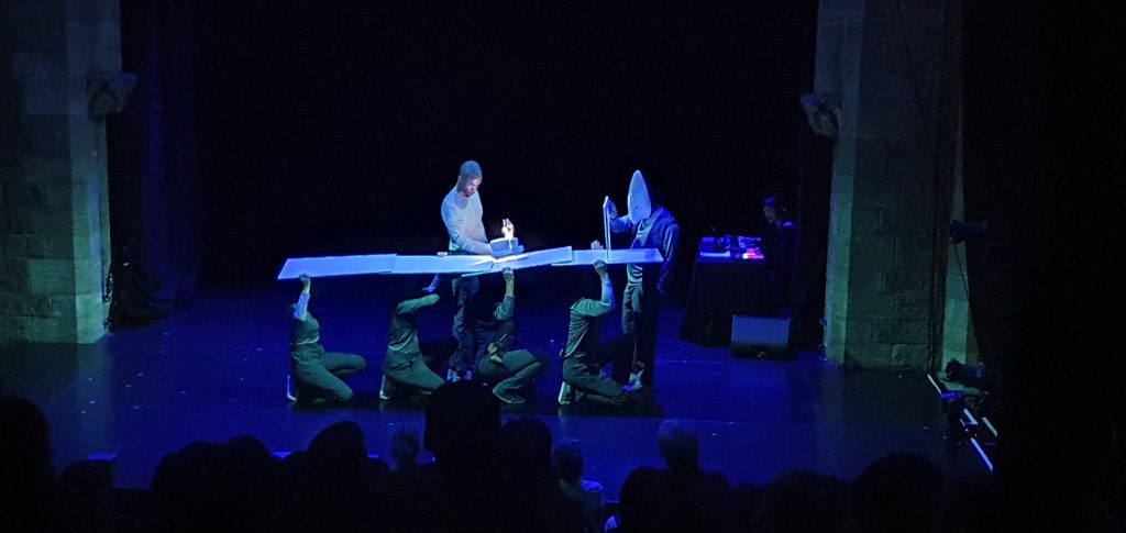 In front of a theatre audience, four performers hold white boards to form a road, along which another performer pushed a model van, while a masked figure staffs a checkpoint.