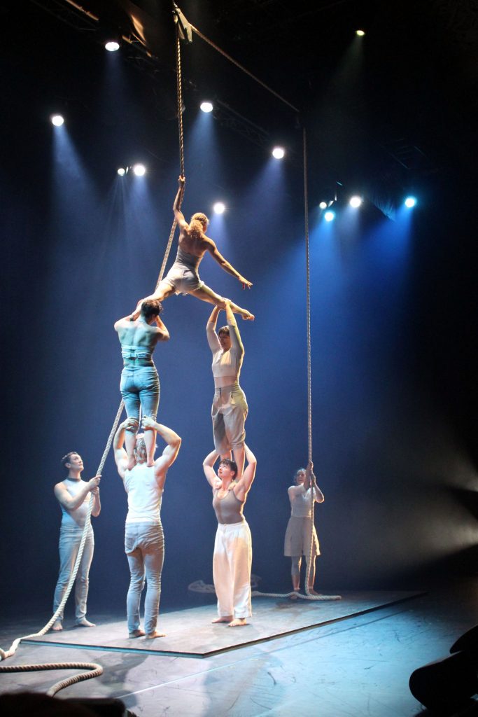 Seven acrobatic performers contribute to a human tower, with one performer doing the splits across the shoulders of two other performers, themselves stood on the shoulders of two more below them.