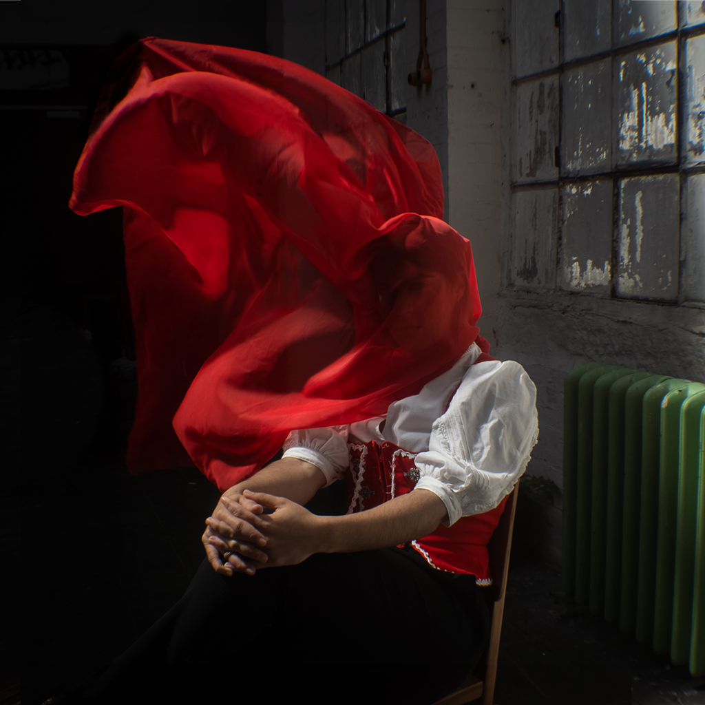 A woman in traditional German dress is obscured by a red scarf violently blown across her face.
